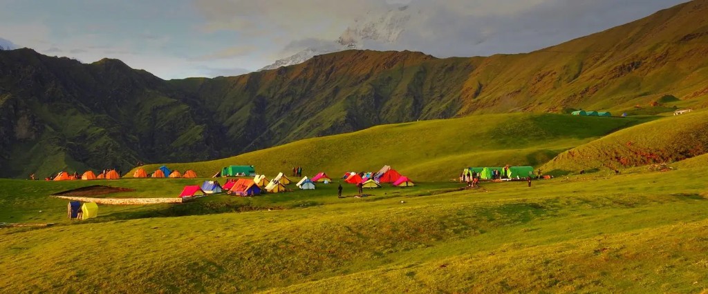 Colorful trekking tents across green rolling meadows at a high-altitude campsite, distant snow peak at golden hour