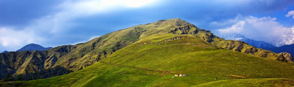 Panoramic lush green rolling meadows of Dayara Bugyal with distant snow-capped Himalayan peaks under a cloudy blue sky