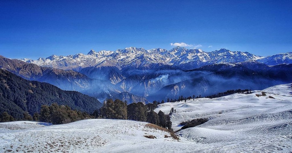 Panoramic view of snow-covered Dayara Bugyal meadow with jagged snow-capped Himalayan peaks under a clear blue sky