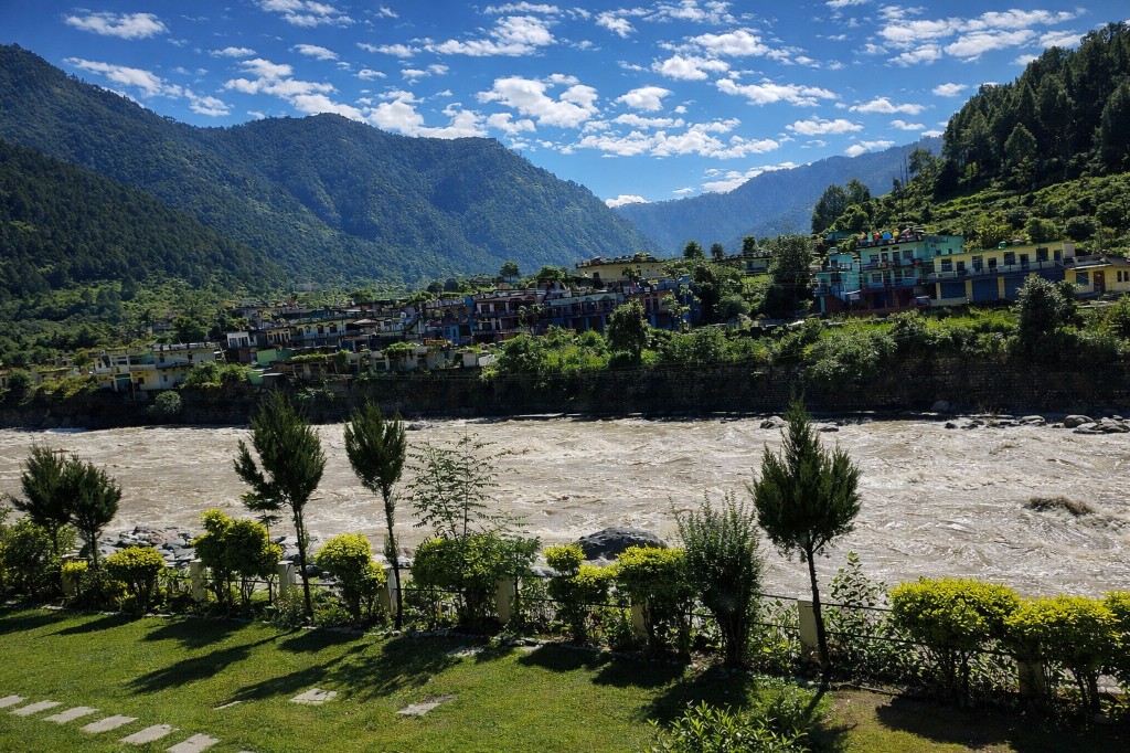 Panoramic view from Shiv Parivar Resort in Uttarkashi — lawn, Himalayan river, hillside village, and mountains